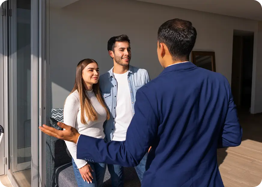 An insurance broker speaks with a smiling couple inside a bright, modern condo with floor-to-ceiling windows.