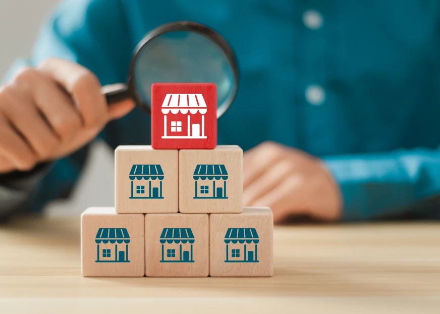 Wooden blocks with small store icons stacked in a pyramid, with a red store block on top and a person holding a magnifying glass in the background.