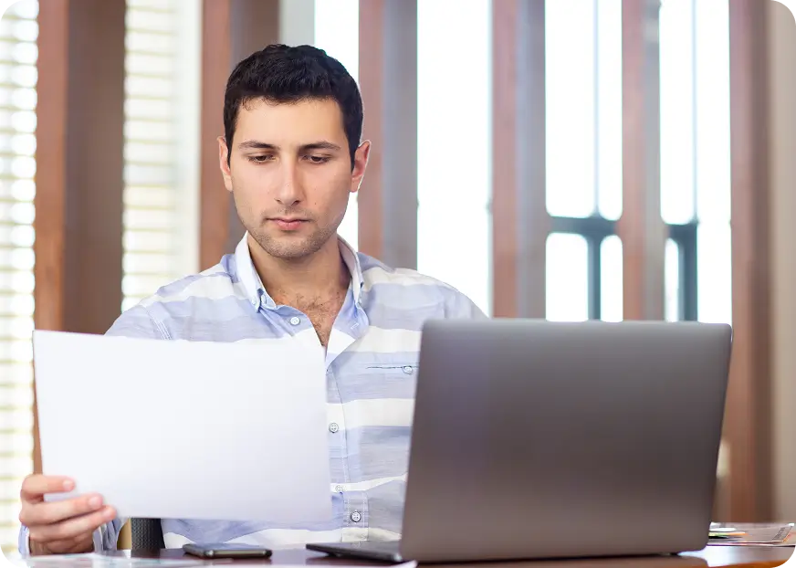 A man reviews insurance documents while sitting at a desk with an open laptop in a modern home office.