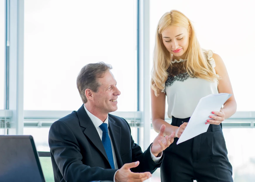 A businesswoman holding a document and speaking with a seated businessman in an office setting.
