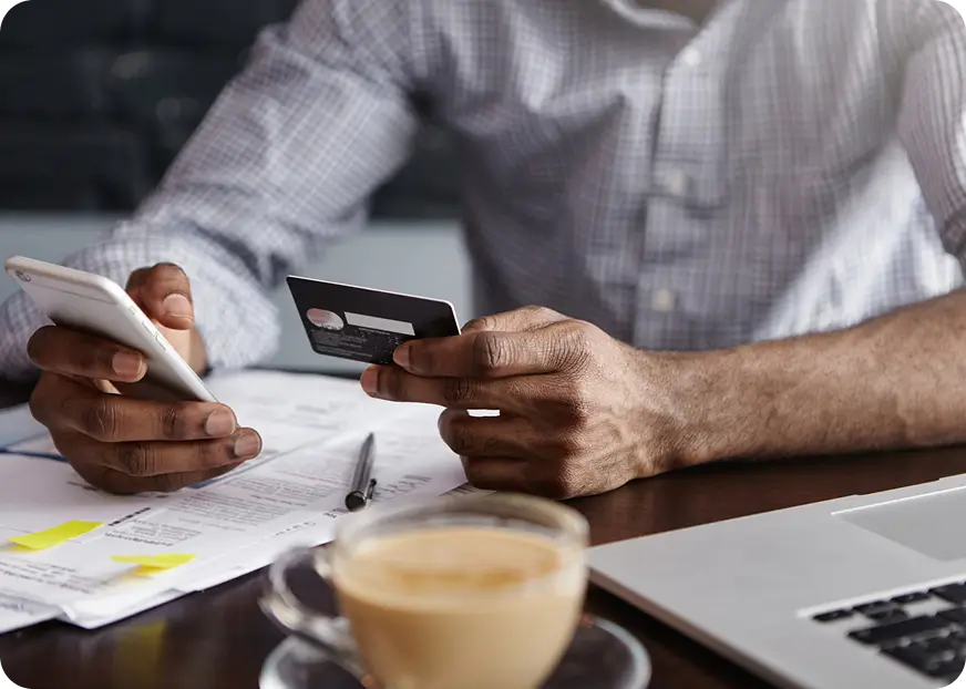 Person using a smartphone and credit card at a desk to complete an online insurance policy payment with documents and laptop nearby